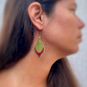 Puede incluir: Un solo pendiente con un diseño tejido verde y rojo. El pendiente tiene forma de diamante con un marco de metal dorado. La parte tejida del pendiente es verde en la parte superior y pasa a rojo en la parte inferior.