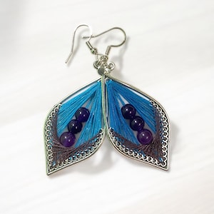 May include: A pair of handmade earrings with a leaf-like design. The earrings feature a silver frame, blue thread, and dark purple beads. The edges are detailed with a silver trim, and the earrings have a hook closure.
