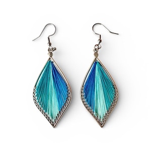 May include: A pair of handmade earrings with a silver-colored metal frame. The earrings feature a teardrop shape filled with blue and teal thread. The edges of the frame are decorated with small, silver-colored beads. The earrings have a hook closure.