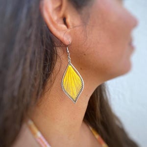 Può includere: Un singolo orecchino di filo giallo con una montatura color argento. L'orecchino ha una forma a goccia con un gancio argentato. I fili gialli sono strettamente intrecciati all'interno della montatura, creando un aspetto strutturato. L'orecchino è indossato all'orecchio di una persona.