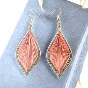 Può includere: Un paio di orecchini dorati a forma di goccia. Gli orecchini presentano un delicato design in filo color corallo intrecciato all'interno di una cornice decorativa. I bordi sono adornati da piccoli accenti circolari.