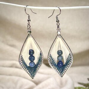 May include: A pair of handcrafted earrings with a teardrop design. Each earring features a silver-colored frame, cream-colored thread, and three blue beads. The bottom of the teardrop is accented with dark teal thread.