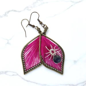 May include: A pair of handmade earrings featuring a leaf-shaped design with bright pink thread. Each earring has a bronze-colored frame and a silver-colored spider charm with a dark gray stone. The earrings have a hook closure.