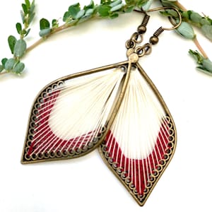 May include: A pair of dangle earrings with a white and red thread design. The earrings are made of brass and have a leaf shape.