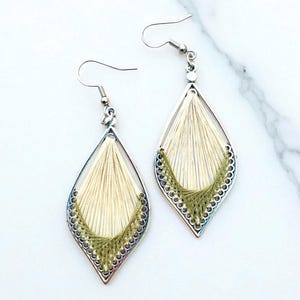 Può includere: Un paio di orecchini fatti a mano a forma di goccia. Gli orecchini presentano una montatura in metallo color argento con un design a filo color crema e dettagli in filo verde oliva. Chiusura a gancio.