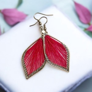 Puede incluir: Pendientes hechos a mano con forma de hoja. Presentan un diseño rojo vibrante tejido dentro de un marco de metal dorado. El marco tiene un delicado borde con cuentas. Los pendientes se exhiben sobre una superficie blanca, con flores rosas en el fondo.