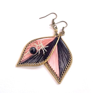 May include: Handmade leaf-shaped earrings with a unique design. The earrings feature a combination of black and pink thread, a bronze-colored frame, and a small silver spider accent with a black bead. The earrings are designed with a hook closure.