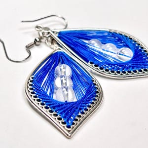 May include: A pair of teardrop-shaped earrings featuring a silver-toned metal frame with a scalloped edge. The center of each earring is filled with vibrant blue thread arranged in a geometric pattern, accented by three clear, spherical beads.
