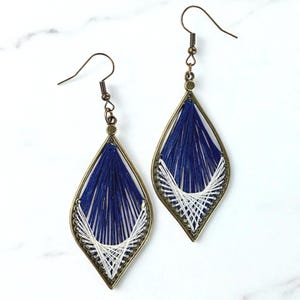 Puede incluir: Un par de pendientes hechos a mano con forma de lágrima. Los pendientes presentan un marco de metal color bronce relleno de hilo azul marino y blanco. El hilo está intrincadamente tejido para crear un patrón único.