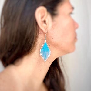 Op de afbeelding: Een paar zilverkleurige oorhangers met een turquoise blauw draadontwerp. De oorbellen hebben een druppelvorm met een delicate, opengewerkte rand. De turquoise draad is strak in het frame geweven.