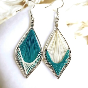 Puede incluir: Un par de pendientes hechos a mano con un marco plateado en forma de lágrima. Un pendiente presenta hilo color turquesa, el otro blanco. Los hilos están intrincadamente tejidos dentro del marco, creando un diseño único y artístico.