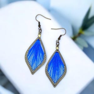 Könnte beinhalten: Ein Paar blaue Fadenohrringe mit einem bronzefarbenen Rahmen. Die Ohrringe haben eine tropfenförmige Form mit einem detaillierten Design. Der blaue Faden ist eng in den Rahmen gewebt und erzeugt ein strukturiertes Aussehen. Die Ohrringe haben einen Hakenverschluss.
