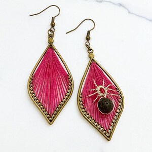 May include: A pair of dangle earrings with a unique design. Each earring features a teardrop-shaped frame with a hot pink thread filling. One earring has a small spider charm. The frames are outlined with a beaded border and have bronze-colored ear wires.
