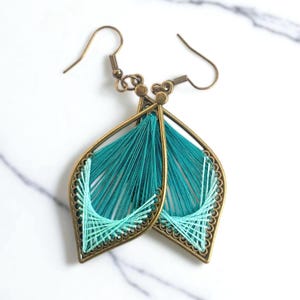 May include: A pair of teal and light teal thread earrings. The earrings have a leaf-like shape with a bronze-colored metal frame. The teal thread is strung across the frame, creating a textured design. The earrings have a hook closure.