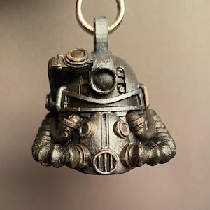 Puede incluir: Un colgante metálico detallado con forma de casco futurista, con intrincados detalles mecánicos y un llavero plateado. El casco tiene una estética industrial desgastada con remaches visibles y detalles circulares, sugiriendo un tema postapocalíptico.