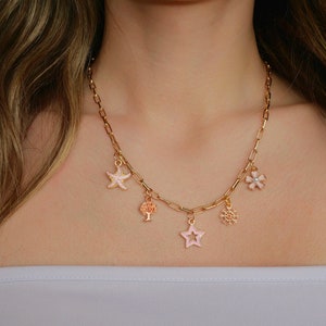 Könnte beinhalten: Eine goldene Kettenkette mit fünf Anhängern: eine rosa Blume, ein Stern, ein Baum, ein Seestern und ein kleines, filigranes Design. Die Anhänger sind alle in Rosa und Weiß emailliert.