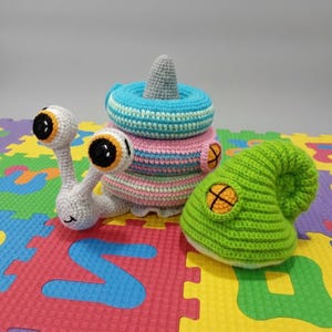 May include: Crocheted toy set featuring a snail with large eyes, a stacking ring toy in blue, pink, and white, and a green hat with an orange cross design. The toys are handmade and suitable for children.