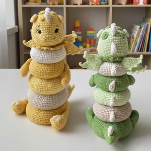 May include: Two crocheted dragon stacking toys. One is yellow with white and tan accents, the other is green with white and tan accents. Each dragon has a head, wings, and a tail. The toys are sitting on a white surface.