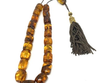 May include: A strand of amber prayer beads with a dark brown tassel and decorative gold-colored metal accents. The beads are oval-shaped and vary in shades of brown and yellow. The beads are strung on a dark brown cord.