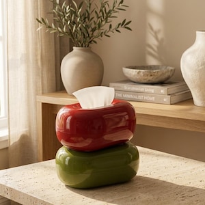 May include: A glossy red tissue box cover stacked on top of a glossy olive green tissue box cover. The red container has a single white tissue protruding from the center slot. Both containers feature a smooth, rounded, pill-shaped design.