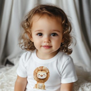 May include: A young child wearing a white t-shirt with a cartoon lion graphic. The lion is brown with a yellow mane and is sitting on its hind legs.