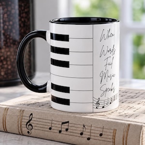 May include: A white ceramic mug with a black handle and interior, featuring a piano keyboard pattern on the side. The text "When Words Fail Music Speaks" is printed in black script, accompanied by a small musical staff and treble clef symbol.