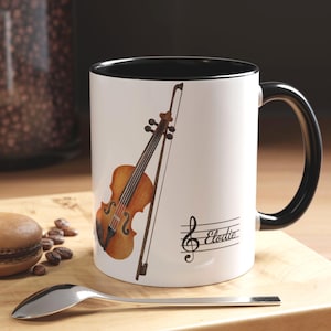 May include: White coffee mug with a black interior and handle. The mug features a watercolor illustration of a violin and bow, with a musical staff and the name "Elodie" printed on the side. The mug is sitting on a wooden surface.