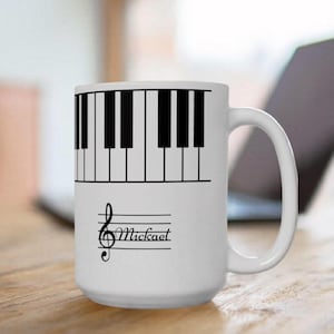 May include: A white ceramic mug featuring a black piano keyboard graphic around the top rim and a black treble clef symbol with the name Mickael written in cursive script on the side, complete with a curved handle and a cylindrical body.