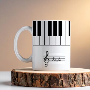 May include: A white ceramic mug featuring a black and white piano keyboard pattern around the top section. Below the keyboard, a black treble clef symbol is printed on a musical staff with the name Kayla written in black script lettering.