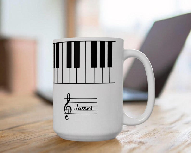 May include: A white ceramic mug featuring a black piano keyboard graphic wrapping around the upper portion. Below the keyboard, a black treble clef symbol is printed next to the name James written in a cursive script on a musical staff.