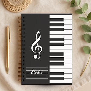 May include: Black spiral-bound notebook with a piano key design and a treble clef. The notebook has the name "Elodie" printed on it. A gold pen and a woven coaster are next to the notebook.