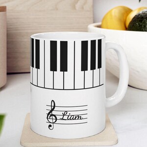 May include: A white ceramic mug featuring a black and white piano keyboard pattern around the top half. The lower half displays a black musical treble clef symbol followed by the name Liam written in a cursive script on a horizontal staff line.