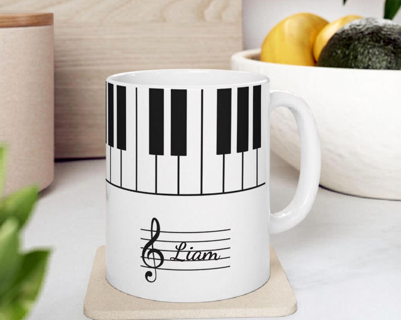 May include: A white ceramic mug featuring a black and white piano keyboard pattern around the top half. The lower half displays a black musical treble clef symbol followed by the name Liam written in a cursive script on a horizontal staff line.