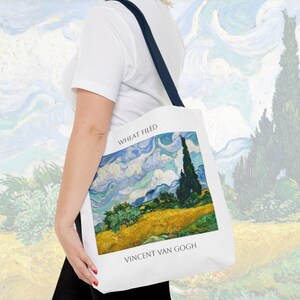 K&ouml;nnte beinhalten: Eine wei&szlig;e Einkaufstasche mit einem blauen und gelben Gem&auml;lde eines Weizenfeldes mit einer Zypresse. Der Text "Wheat Field" und "Vincent van Gogh" ist auf der Tasche aufgedruckt.