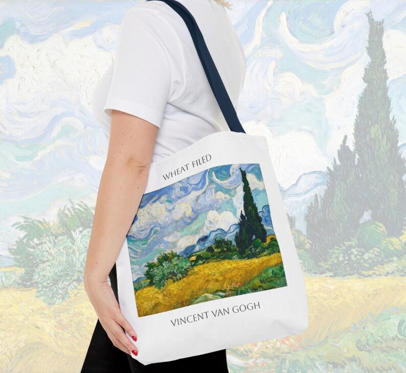 K&ouml;nnte beinhalten: Eine wei&szlig;e Einkaufstasche mit einem blauen und gelben Gem&auml;lde eines Weizenfeldes mit einer Zypresse. Der Text "Wheat Field" und "Vincent van Gogh" ist auf der Tasche aufgedruckt.