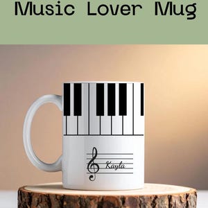 May include: A white ceramic mug featuring a black and white piano keyboard pattern around the top rim. Below the keys, a black musical staff with a treble clef and a customizable name written in cursive script is printed on the front surface.