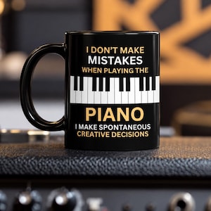 May include: A black ceramic mug featuring a white piano keyboard graphic in the center. The text reads: I DON'T MAKE MISTAKES WHEN PLAYING THE PIANO I MAKE SPONTANEOUS CREATIVE DECISIONS. The mug has a curved handle and a smooth, glossy finish.