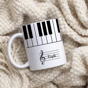 May include: A white ceramic mug featuring a black and white piano key pattern around the top rim. Below the keys, a black musical treble clef symbol is printed alongside the name Kayla in a cursive script font on a horizontal staff line.