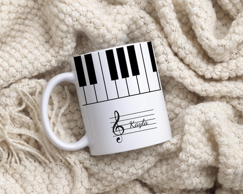 May include: A white ceramic mug featuring a black and white piano key pattern around the top rim. Below the keys, a black musical treble clef symbol is printed alongside the name Kayla in a cursive script font on a horizontal staff line.
