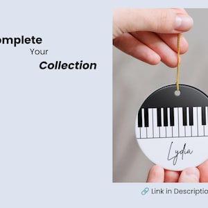 May include: A round, flat, white and black ceramic ornament featuring a piano keyboard graphic and the name Lydia in black script. The ornament hangs from a gold string and includes the text Complete Your Collection and Link in Description.