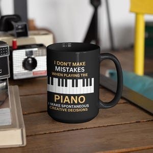 May include: Black coffee mug with white text that reads "I don't make mistakes when playing the piano. I make spontaneous creative decisions."  The text is in a gold color.  There is a black and white image of a piano keyboard below the text.