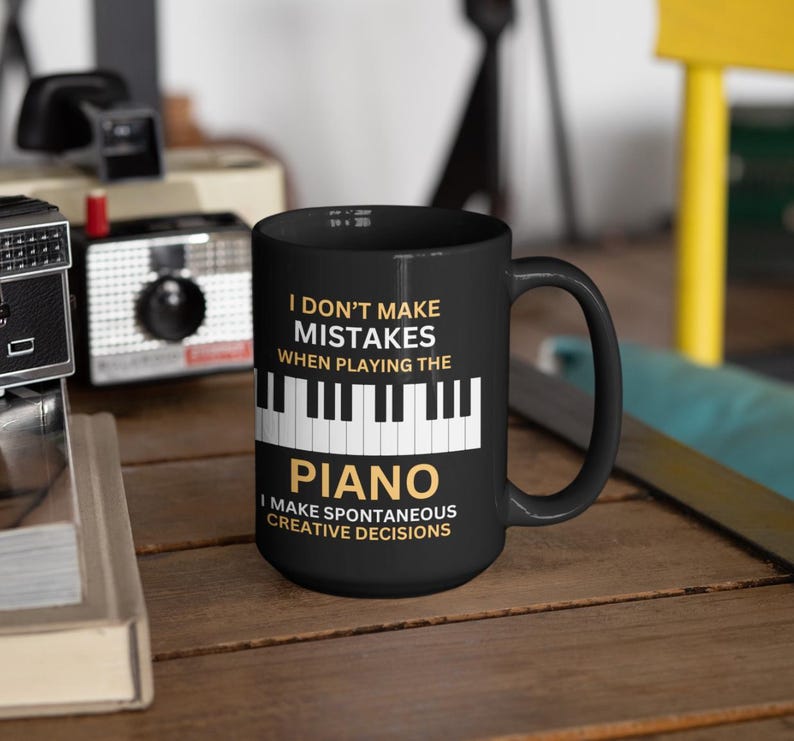 May include: Black coffee mug with white text that reads "I don't make mistakes when playing the piano. I make spontaneous creative decisions."  The text is in a gold color.  There is a black and white image of a piano keyboard below the text.
