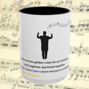 May include: A white ceramic mug with a black interior rim featuring a black silhouette of an orchestra conductor holding a baton with musical notes above. The text reads: There are two golden rules for an orchestra start together and finish together. The public doesn't give a damn what goes on in between - Thomas Beecham.