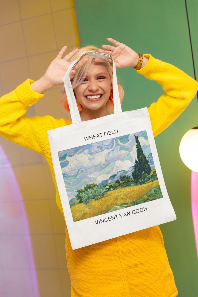 K&ouml;nnte beinhalten: Eine wei&szlig;e Einkaufstasche mit einem Gem&auml;lde eines Weizenfeldes von Vincent van Gogh. Das Gem&auml;lde tr&auml;gt den Titel "Weizenfeld" und der Name des K&uuml;nstlers ist unter dem Bild gedruckt.