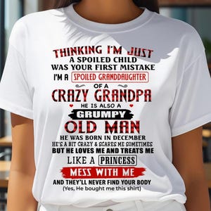 May include: White t-shirt with a humorous message in red and black plaid and bold text. The text reads "Thinking I'm Just a Spoiled Child Was Your First Mistake, I'm a Spoiled Granddaughter of a Crazy Grandpa".