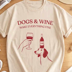 May include: Beige t-shirt with rolled sleeves featuring a red graphic of a hand holding a wine glass, a wine bottle, and a dog. The text "DOGS & WINE MAKE EVERYTHING FINE" is printed above the graphic.