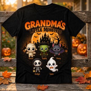 May include: A black t-shirt featuring the orange text "GRANDMA'S LITTLE MONSTERS" above a Halloween scene with a haunted house, bats, and five cartoon monsters: a mummy, Frankenstein, a bat, a skeleton, and a ghost with names below each.