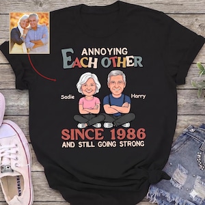 Può includere: T-shirt nero con la scritta "Annoying Each Other Since 1986 and Still Going Strong". Presenta illustrazioni di cartoni animati di una coppia, Sadie e Harry, con una foto della coppia in alto a sinistra.