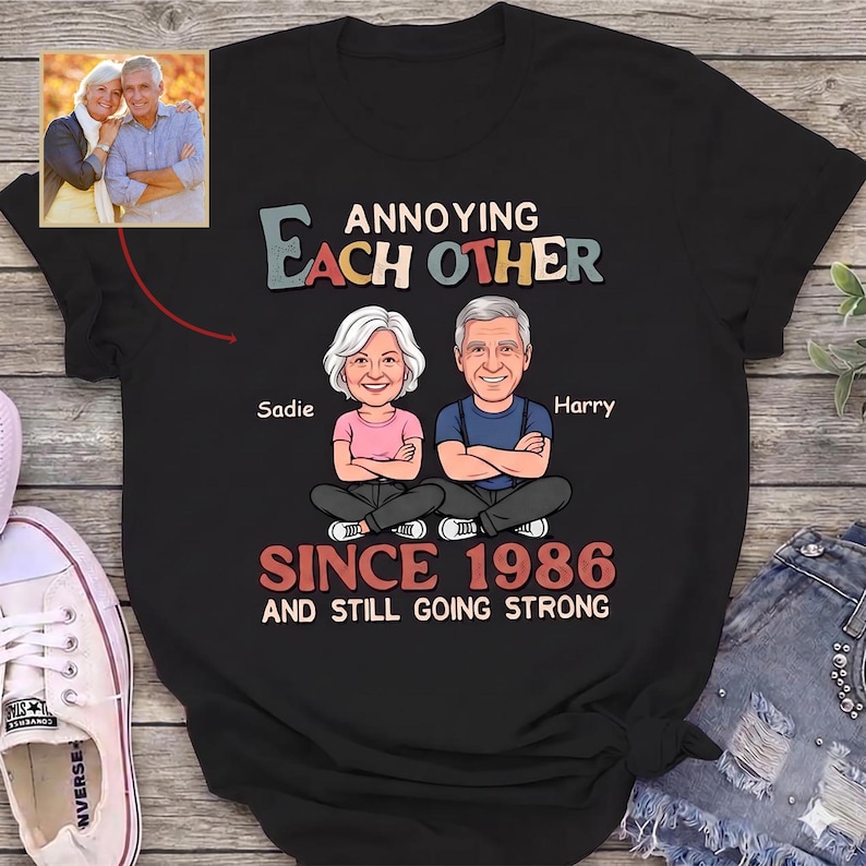 Puede incluir: Camiseta negra con el texto "Annoying Each Other Since 1986 and Still Going Strong". Presenta ilustraciones de dibujos animados de una pareja, Sadie y Harry, con una foto de la pareja en la esquina superior izquierda.