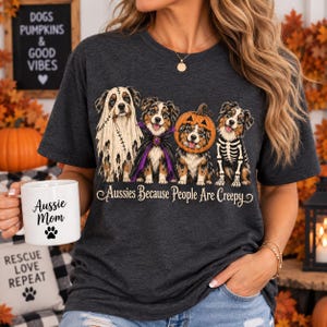 Puede incluir: Una camiseta gris oscuro con un gráfico de cuatro perros pastores australianos con disfraces de Halloween, incluyendo un fantasma, un vampiro, una calabaza y un esqueleto. El texto debajo de los perros dice "Aussies Because People Are Creepy."